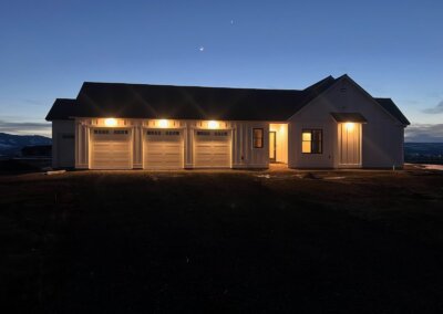 Detached three-car garage with dark-colored garage doors at dusk, with a matching home visible in the background and exterior lighting highlighting the doors.