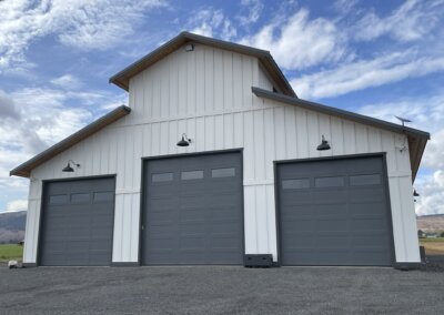 Large shop-style building with three dark gray garage doors, white siding, and black exterior lights, photographed in daylight on a rural property.